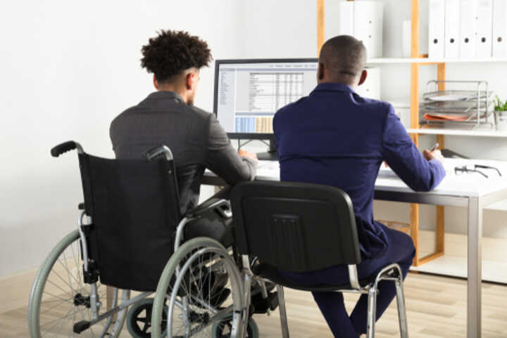 Rear view of two colleagues sitting at a desk with a computer; one man is in a wheelchair