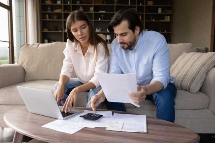 Couple on sofa at home, checking paperwork with a laptop and calculator